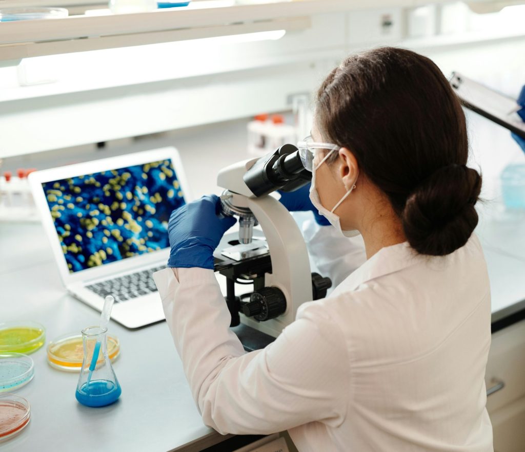 A female scientist in a lab coat examines samples under a microscope for research.