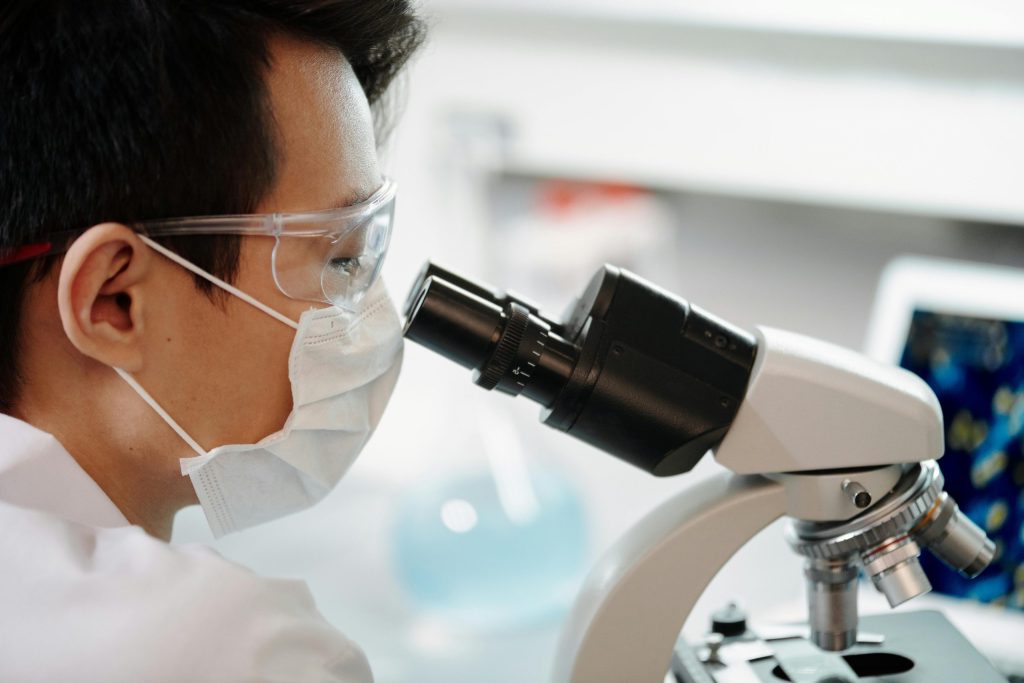 Scientist wearing face mask and goggles examining samples through a microscope in a lab.